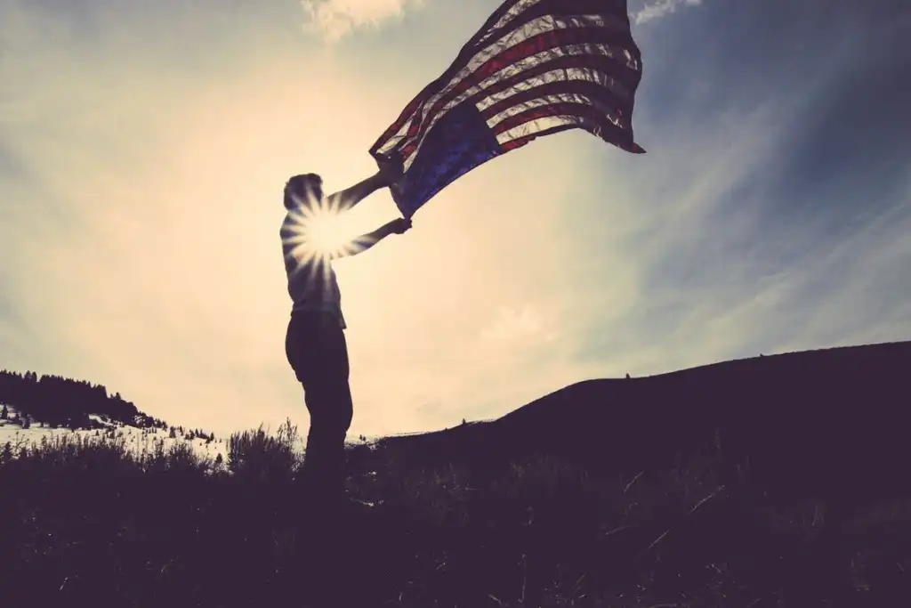 Veteran holding American flag at sunset with light shining through, symbolizing service and patriotism